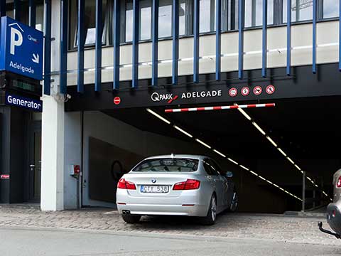 Q-Park Adelgade p-hus indkørsel. Skilte og dele af bygningen synlig. Sølv bil kører ind.