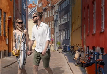 A woman and a man holding hands, walking along a cobblestone street with colourful buildings – 24 Hours in the City