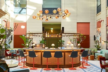 Stylish, modern bar area featuring a circular bar counter, bar stools, and decorative lighting, surrounded by bottles and plants – Parking at 25hours Hotel in Copenhagen