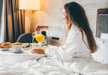 Kvinde der sidder i hotelseng og spiser lækker morgenmad med appelsinjuice, brød og frugt - Hotelparkering Q-Park