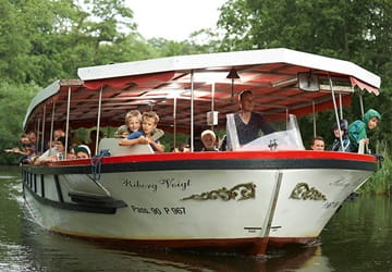 Åfartsbåd sejler på en å omgivet af grøn natur, med voksne og børn ombord under en skyggefuld kaleche - Parkering Odense Aafart