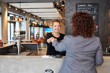 Hotel reception and café counter where a receptionist serves a customer, with shelves of bottles, hanging lights, and a menu board in the background - Parking Zleep Hotel Aalborg