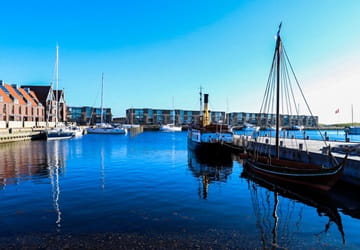 Sailboats and traditional wooden vessels are moored in a marina in Frederikssund. The calm water reflects the boats, quays and waterfront buildings.